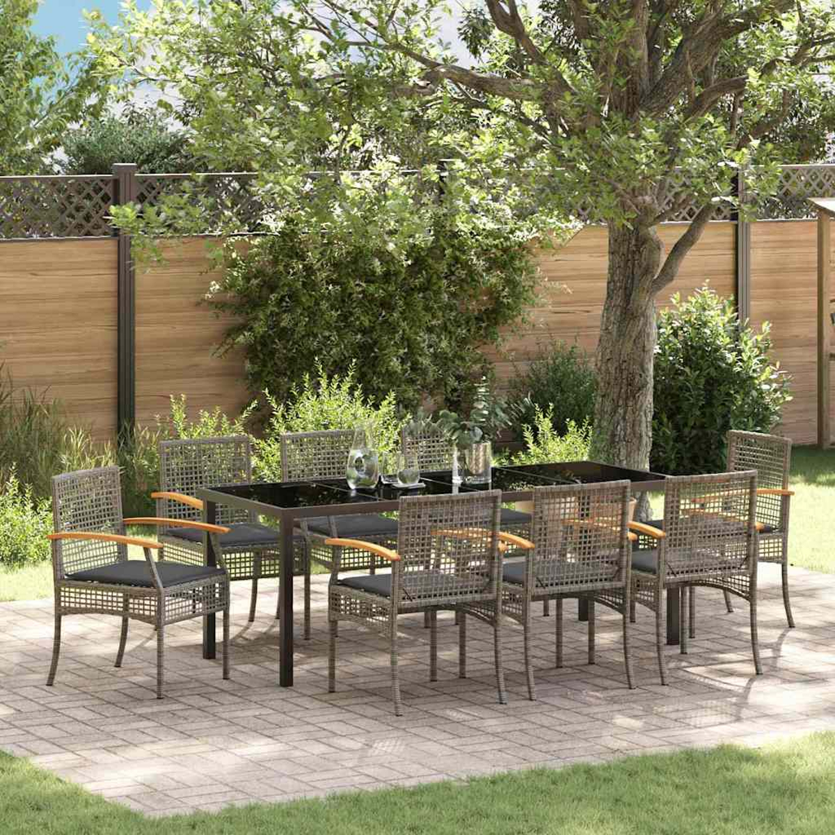 Set da pranzo all'aperto: un tavolo nero, otto sedie, su un patio, sotto un albero. Recinzione in legno e vegetazione sullo sfondo.