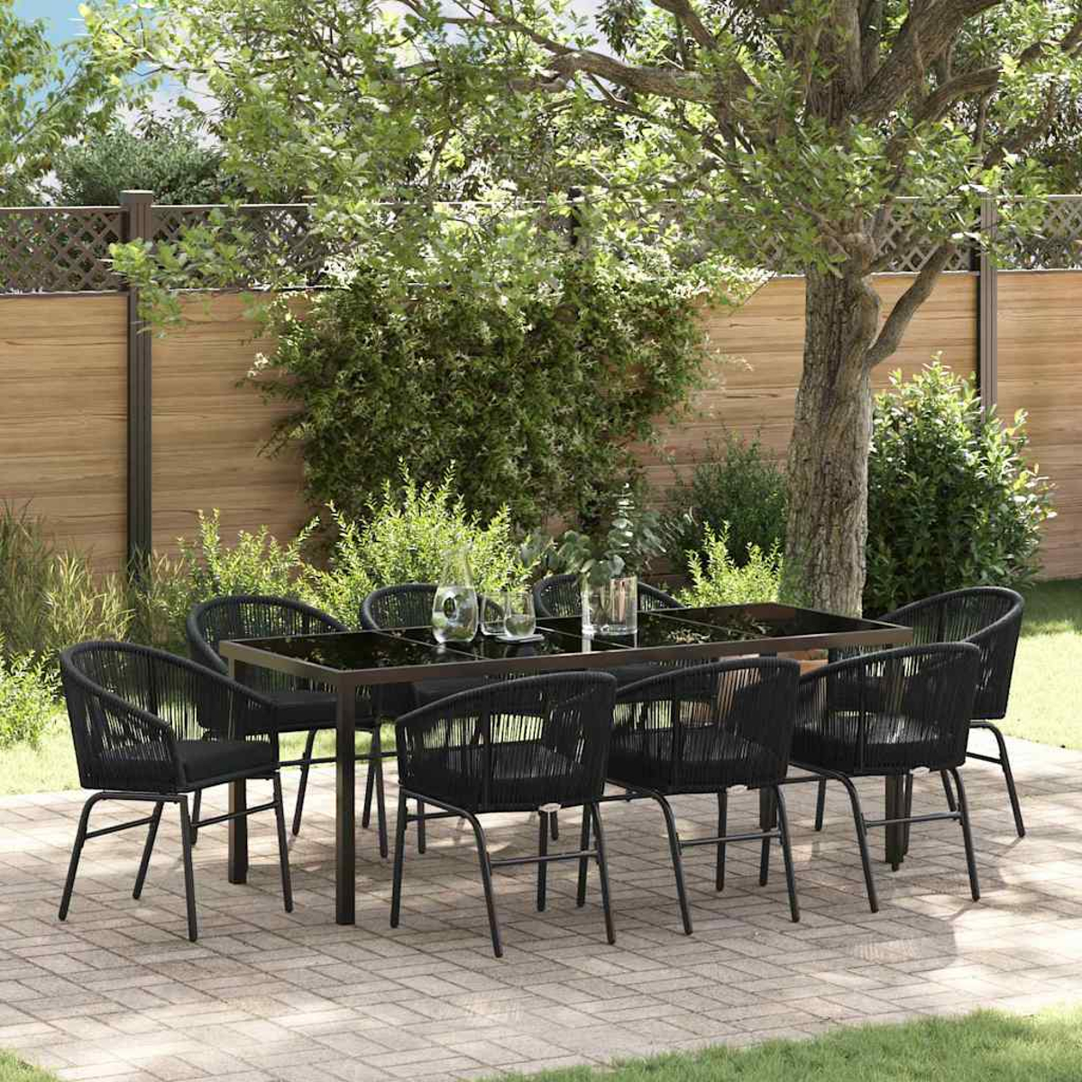 Juego de comedor al aire libre con sillas y mesa negras. La escena incluye una valla de madera, plantas verdes y un árbol.