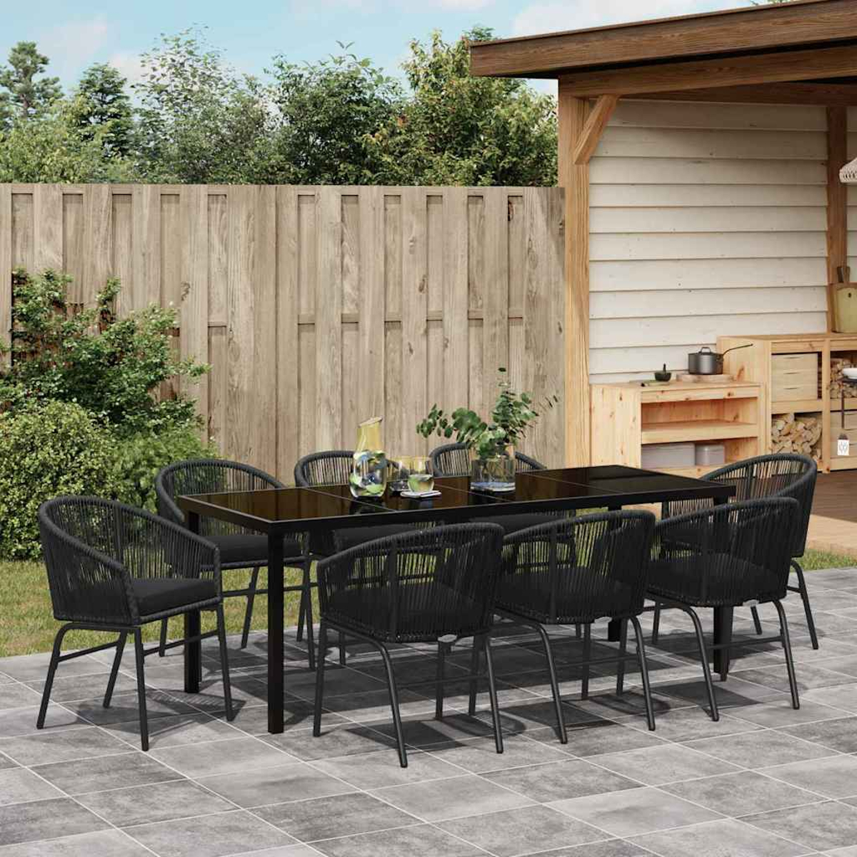 Juego de comedor al aire libre: mesa y sillas negras en el patio. Valla y estructura de madera al fondo. Plantas y bebidas en la mesa.