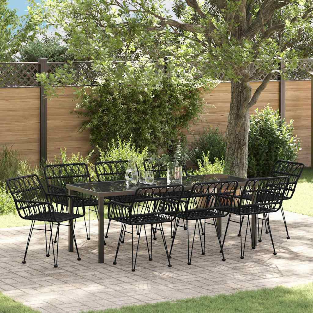 Juego de comedor al aire libre con sillas negras y una mesa con tapa de vidrio en un patio de ladrillos. Vegetación exuberante y una valla de madera en el fondo.