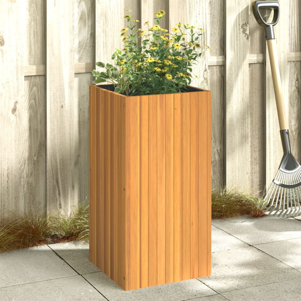Ein hoher Holzpflanzer mit gelben Blumen. Eine Harke lehnt an einem Holzzaun. Der Boden ist gepflastert.