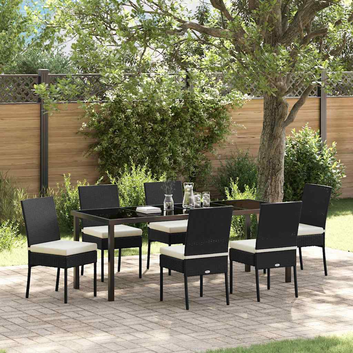 Essgruppe im Freien: schwarzer Tisch und Stühle mit cremefarbenen Kissen, auf einer Ziegelterrasse. Viel Grün und Bäume im Hintergrund.