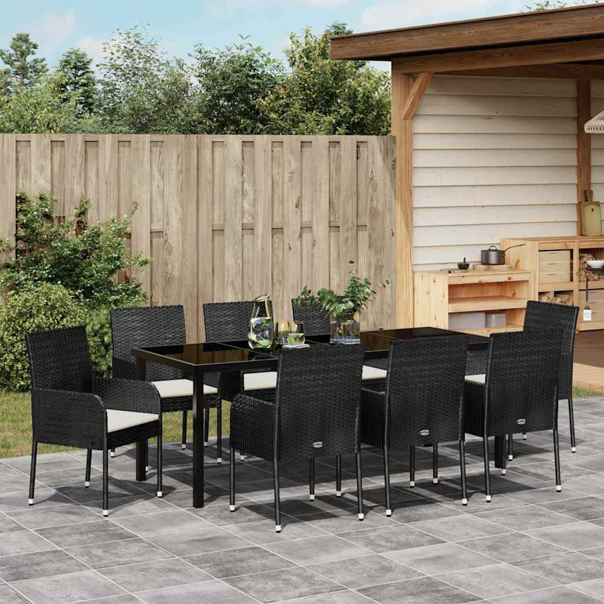 Essgruppe im Freien. Schwarze Korbstühle und Tisch auf einer gefliesten Terrasse. Holzzaun und -struktur im Hintergrund. Pflanzen und Getränke auf dem Tisch.
