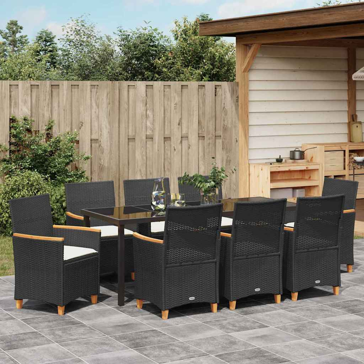 Essgruppe für die Terrasse. Schwarze Korbstühle und ein Tisch. Holzzaun und Struktur im Hintergrund. Grüne Büsche und Bäume.