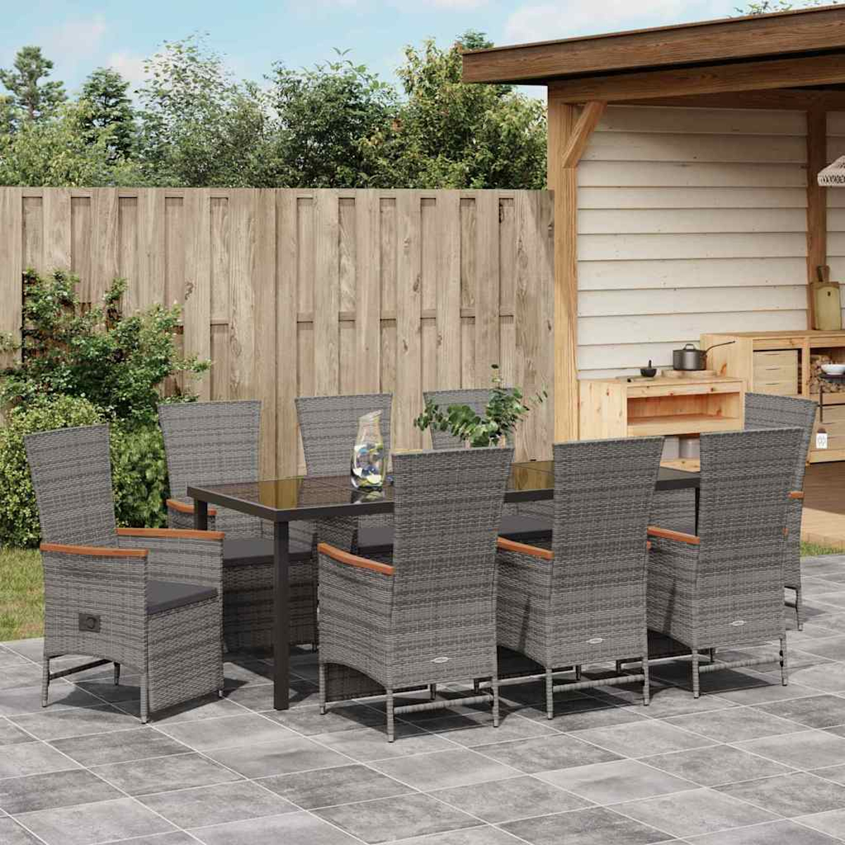 Essgruppe im Freien. Graue Korbstühle und ein Tisch stehen auf einer Terrasse. Holzzaun und ein überdachter Bereich sind im Hintergrund.