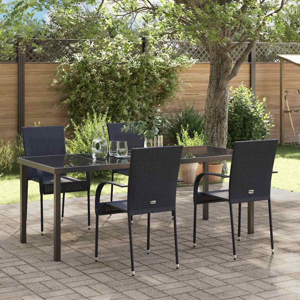 Eine Terrasse mit einem Tisch und vier schwarzen Stühlen. Eine Kanne und Gläser stehen auf dem Tisch. Im Hintergrund grüne Bäume und ein Zaun.