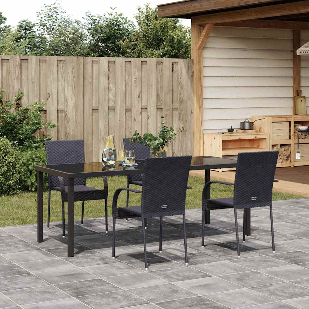 Szene auf der Terrasse mit Tisch und Stühlen. Ein Krug, Gläser und Pflanzen stehen auf dem Tisch. Im Hintergrund ein Holzzaun und eine Struktur.