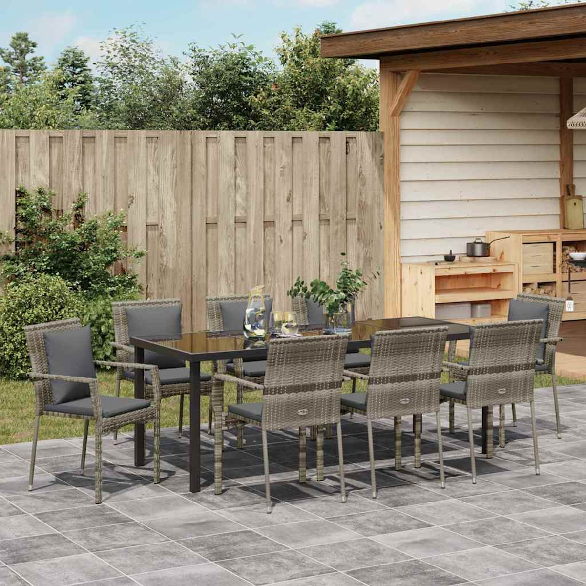 Essgruppe im Freien: Tisch mit Glasplatte und acht Stühlen. Terrasse mit Holzzaun, Pflanzen und kleiner Außenküche.