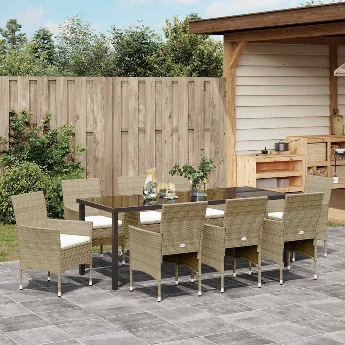Essgruppe im Freien. Ein Tisch mit acht Stühlen auf einer Terrasse. Im Hintergrund ein Holzzaun, Pflanzen und eine kleine Außenküche.