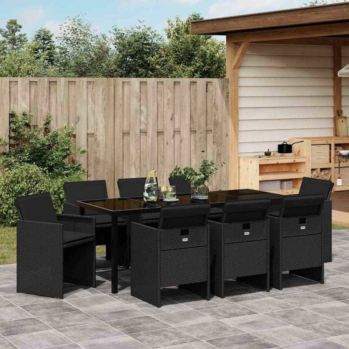 Essgruppe im Freien mit schwarzen Korbstühlen und einem Tisch mit Glasplatte. Auf einer gefliesten Terrasse, mit Holzzaun und Schuppen im Hintergrund.
