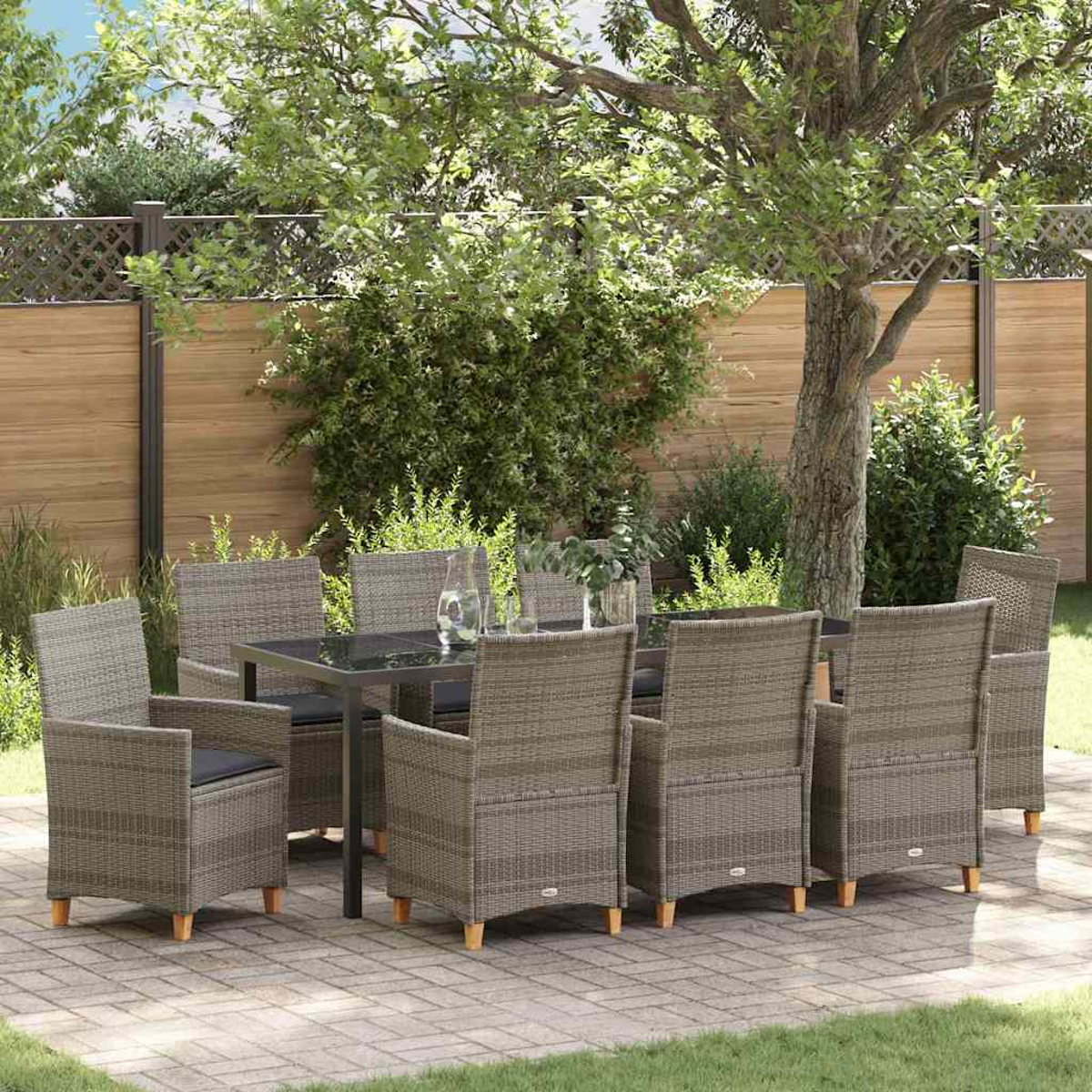 Essgruppe im Freien. Ein Tisch mit Glasplatte und acht Korbstühlen auf einer Terrasse mit Grün und Holzzaun.
