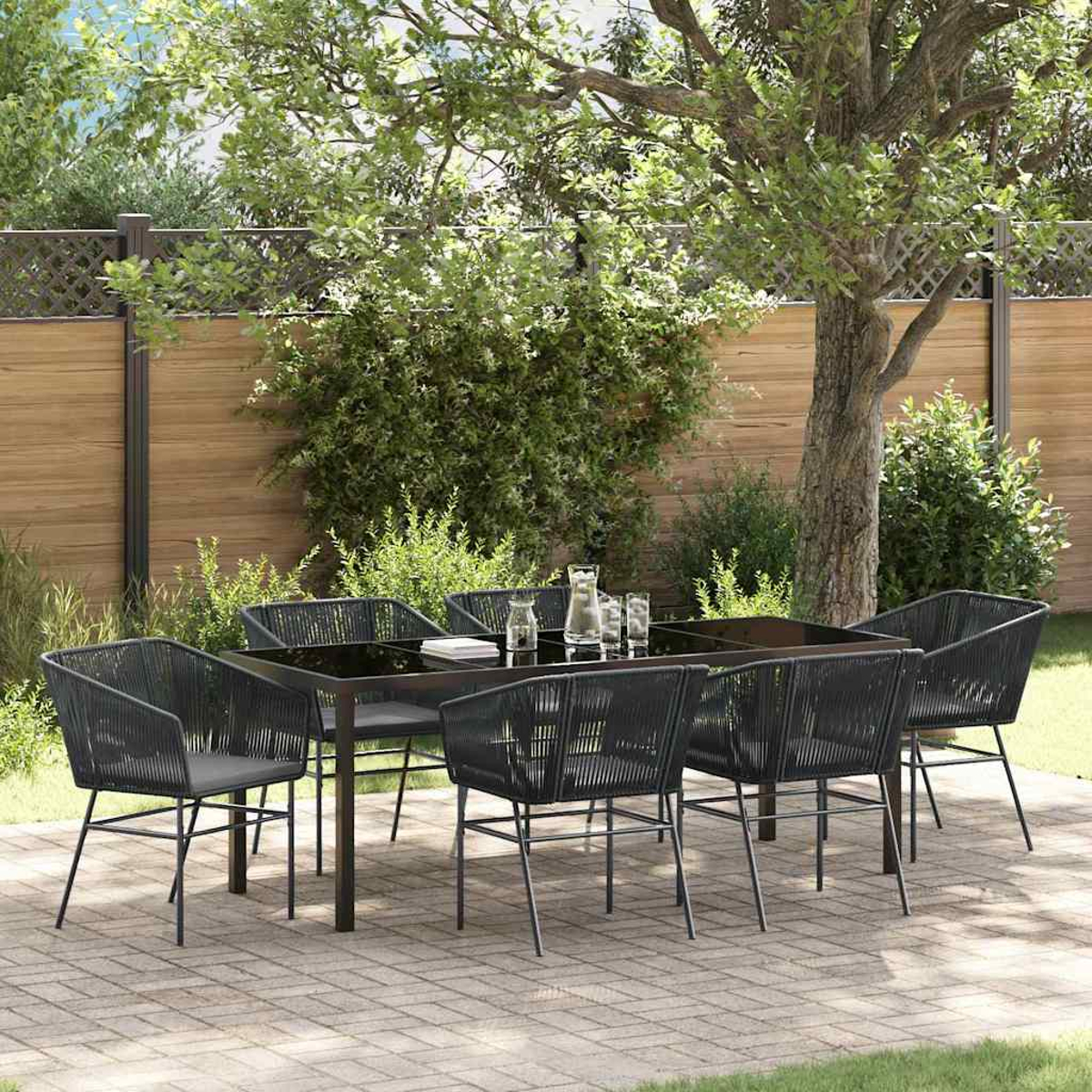 Essgruppe im Freien: schwarzer Tisch, Stühle, Gläser und ein Krug. Auf einer Ziegelterrasse, mit Holzzaun und Bäumen im Hintergrund.