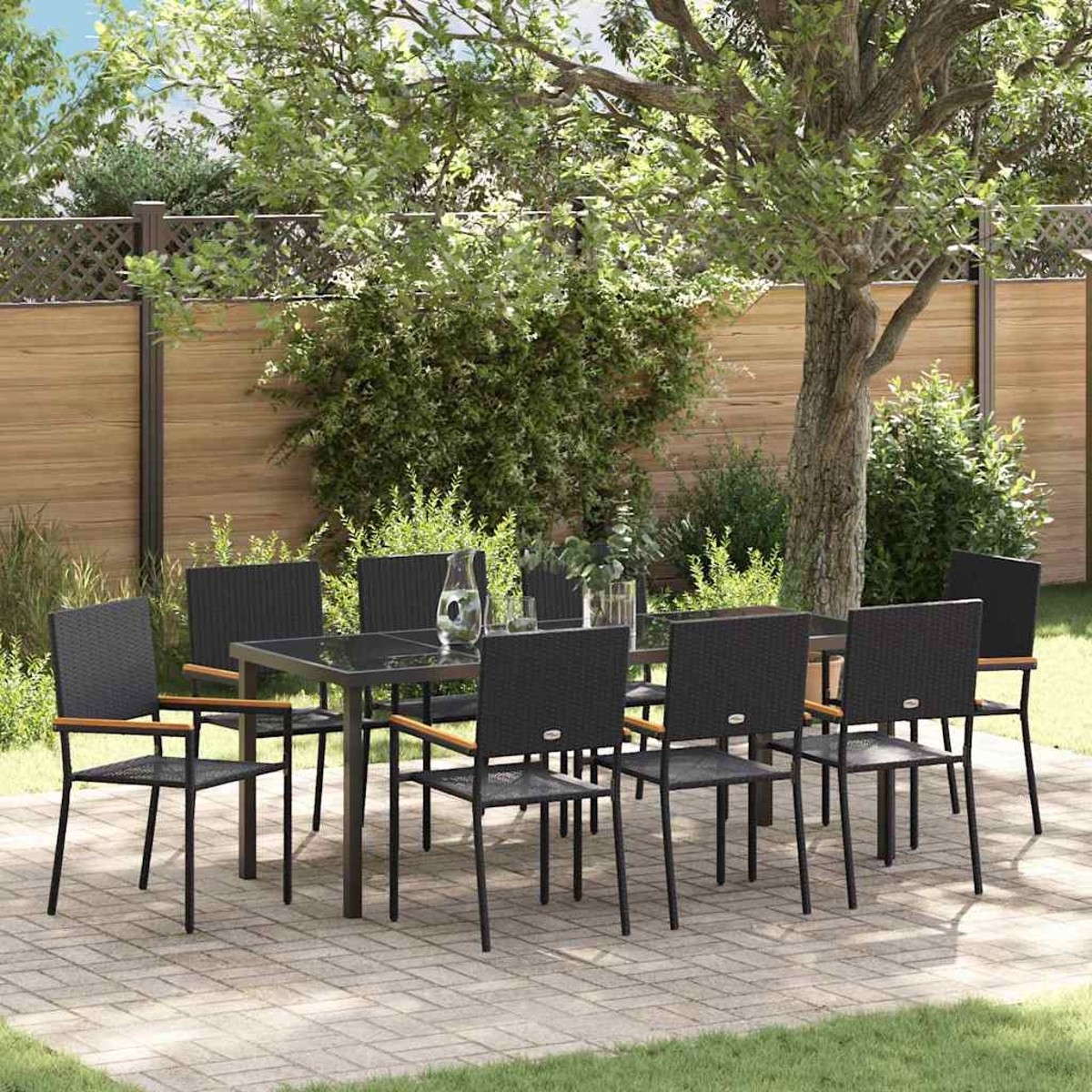 Juego de comedor al aire libre: sillas negras, mesa de vidrio en un patio. Césped verde, valla de madera y árboles al fondo.