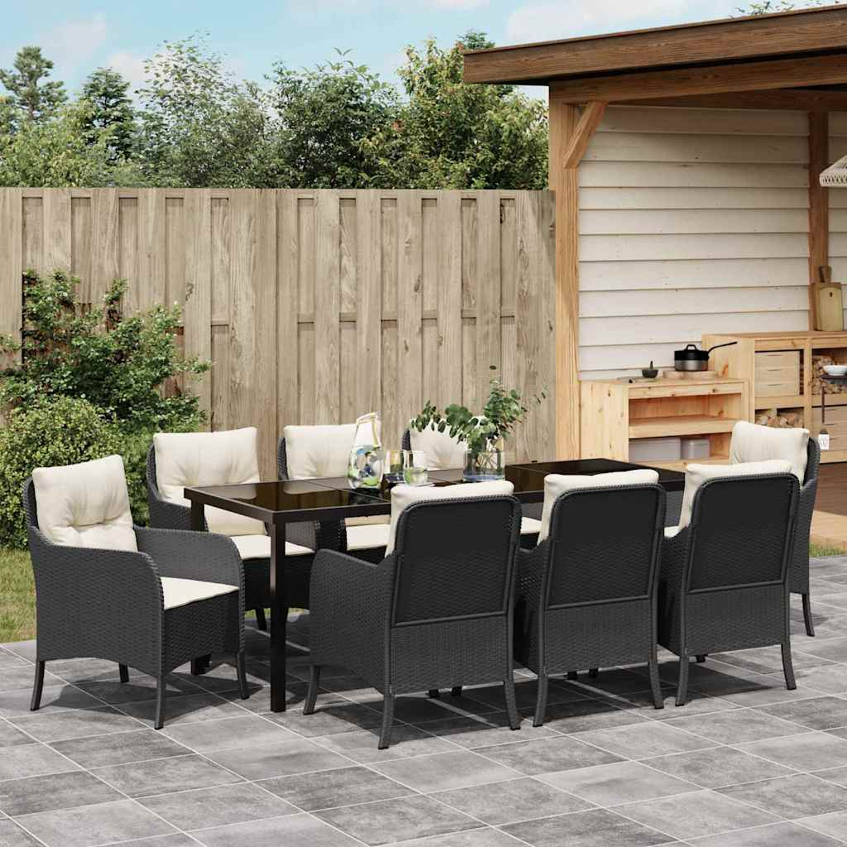 Essgruppe im Freien mit schwarzen Stühlen und einem Tisch mit Glasplatte. Beinhaltet beige Kissen. Auf einer gefliesten Terrasse. Holzzaun und Schuppen im Hintergrund.