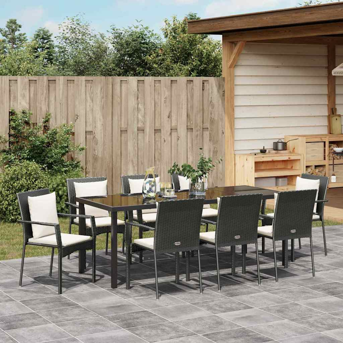 Essgruppe im Freien. Schwarzer Tisch mit Stühlen auf einer Terrasse, mit Holzzaun und kleinem Schuppen im Hintergrund.