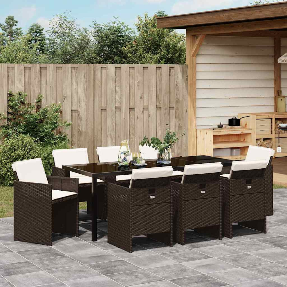 Essgruppe im Freien auf der Terrasse: braune Korbstühle und Tisch mit weißen Kissen. Im Hintergrund ein Holzzaun und ein Schuppen.