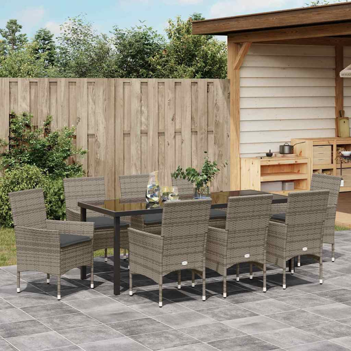 Essgruppe im Freien: Tisch, acht Stühle auf der Terrasse. Holzzaun und Struktur im Hintergrund. Pflanzen und Geschirr auf dem Tisch.