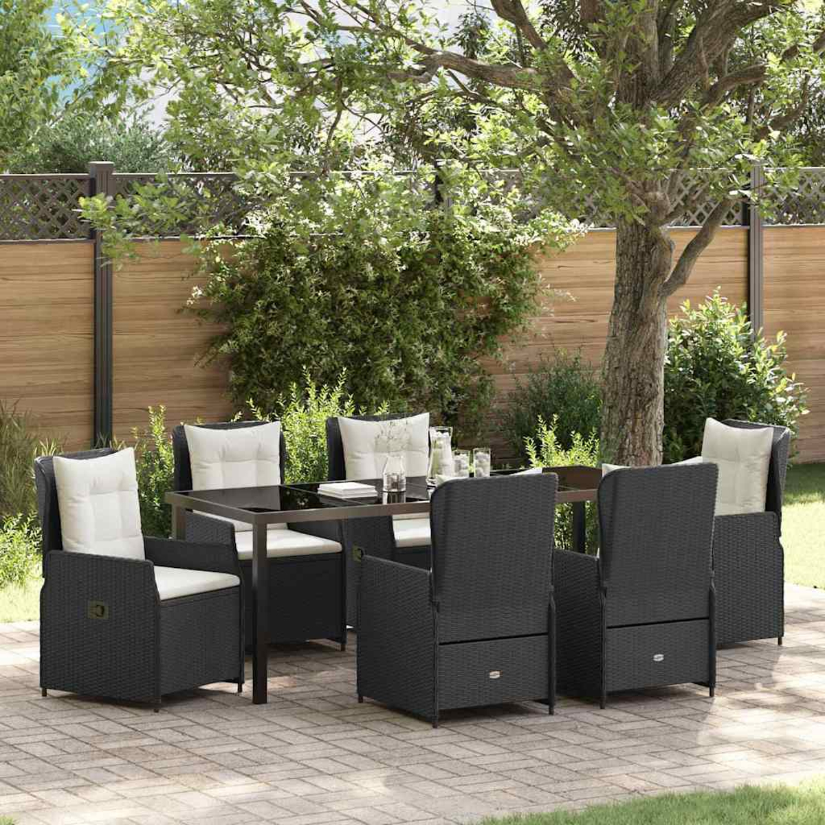 Essgruppe im Freien: schwarze Korbstühle mit weißen Kissen, Tisch mit Glasplatte, in einem Garten, mit Holzzaun und Bäumen.