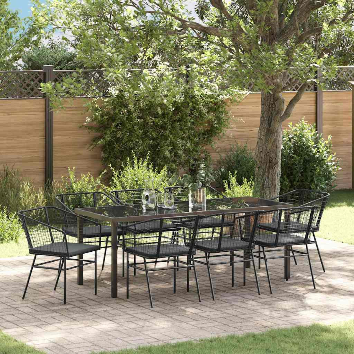 Essgruppe im Freien: Schwarze Metallstühle und -tisch auf einer Terrasse. Grüne Pflanzen, Bäume und Holzzaun im Hintergrund.