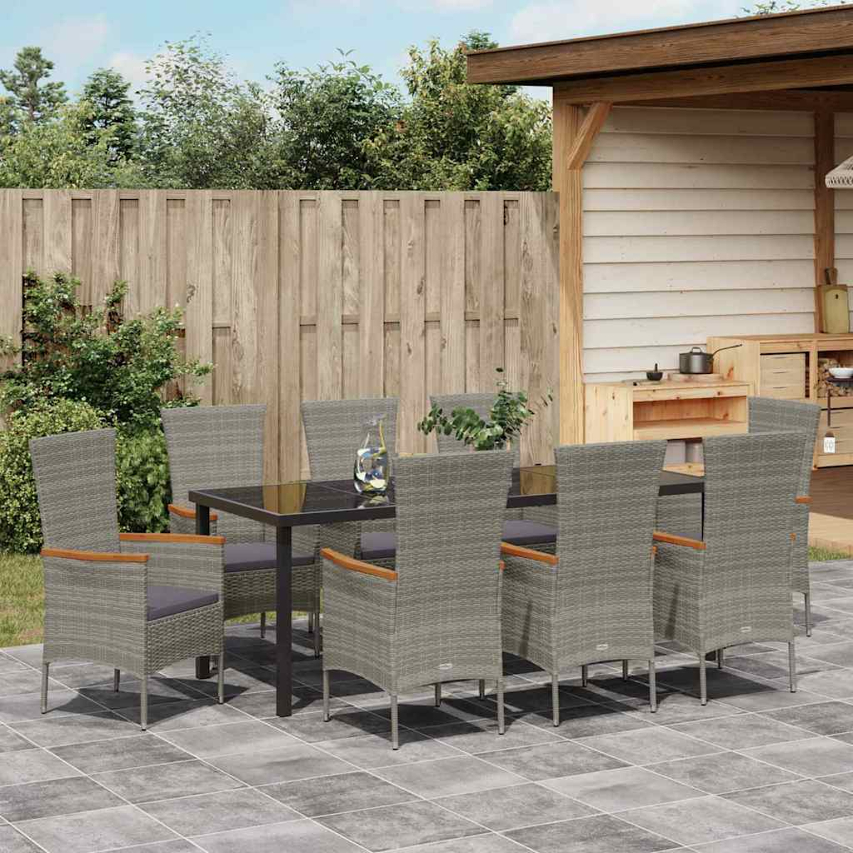 Essgruppe im Freien: graue Korbstühle um einen schwarzen Tisch auf einer gefliesten Terrasse. Holzzaun und Struktur im Hintergrund.
