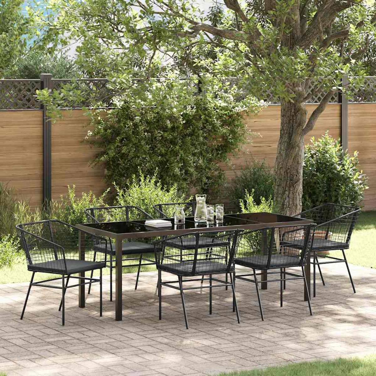 Terrassengarnitur mit schwarzen Metallstühlen, einem Tisch mit Glasplatte und einer Kanne. In einem Garten mit Holzzaun und Bäumen.