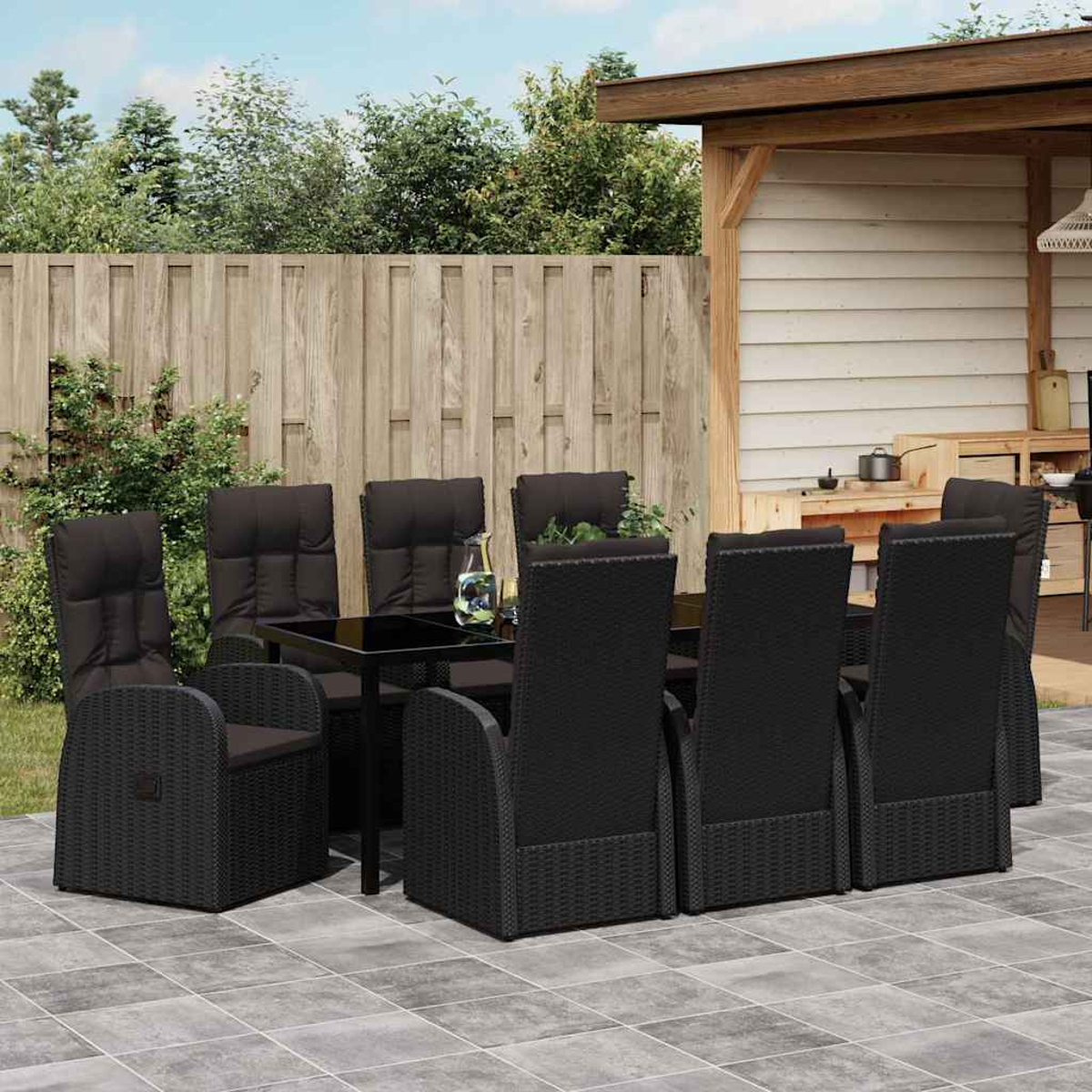 Essgruppe im Freien. Schwarze Korbstühle und Tisch auf der Terrasse. Holzzaun und -struktur im Hintergrund. Pflanzen und Himmel sichtbar.