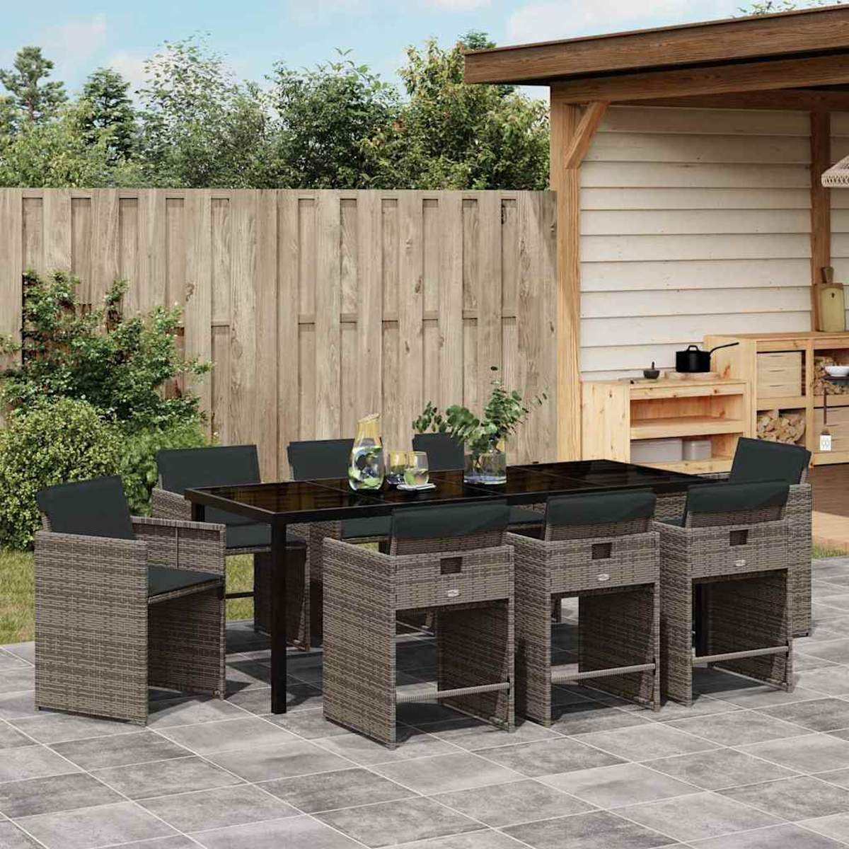 Essgruppe im Freien. Auf einer Terrasse ist ein Tisch mit Stühlen aufgebaut. Der Hintergrund zeigt einen Holzzaun und eine kleine Küche.