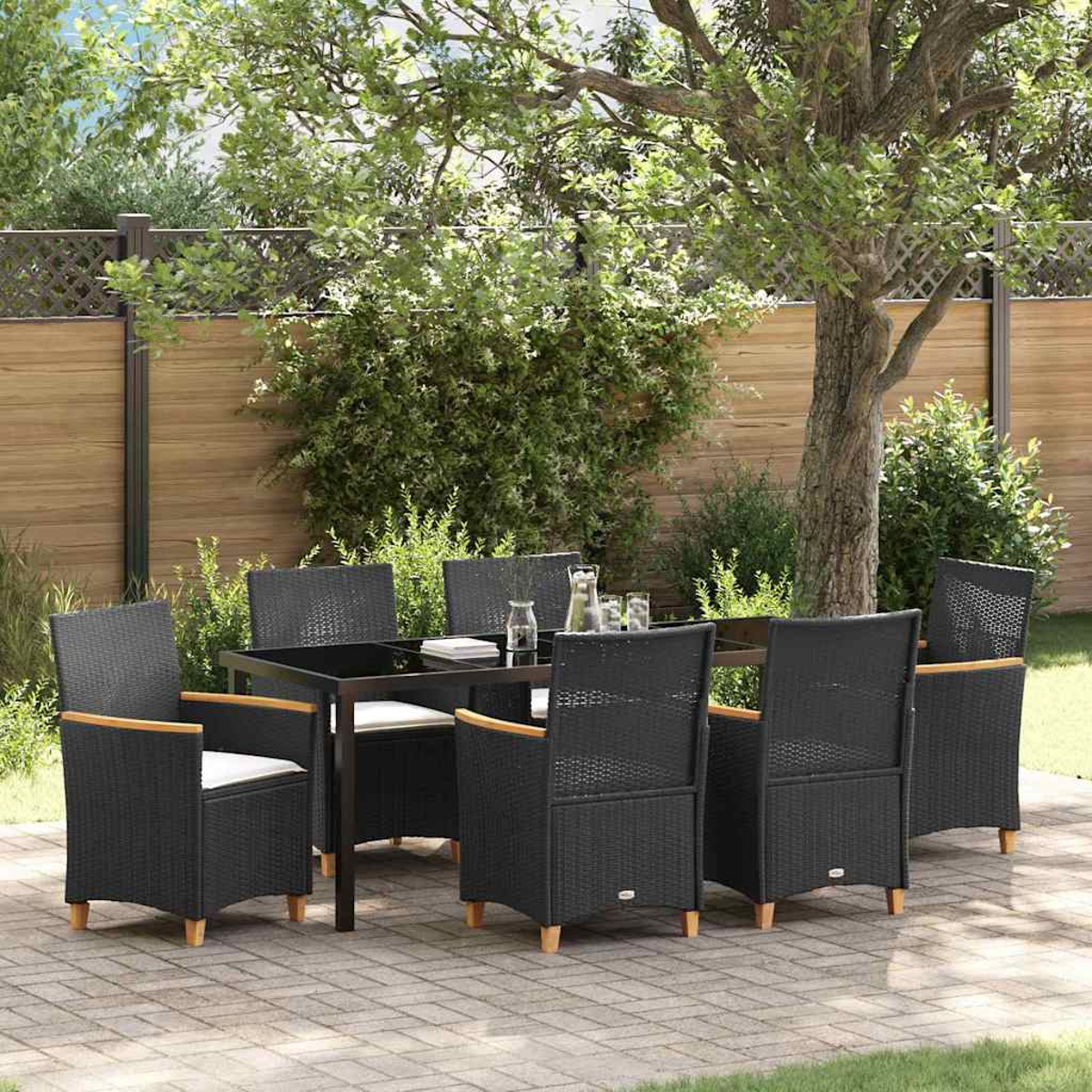 Essgruppe im Freien: schwarze Korbstühle, Tisch mit Glasplatte, in einem Garten. Holzzaun und Bäume im Hintergrund.
