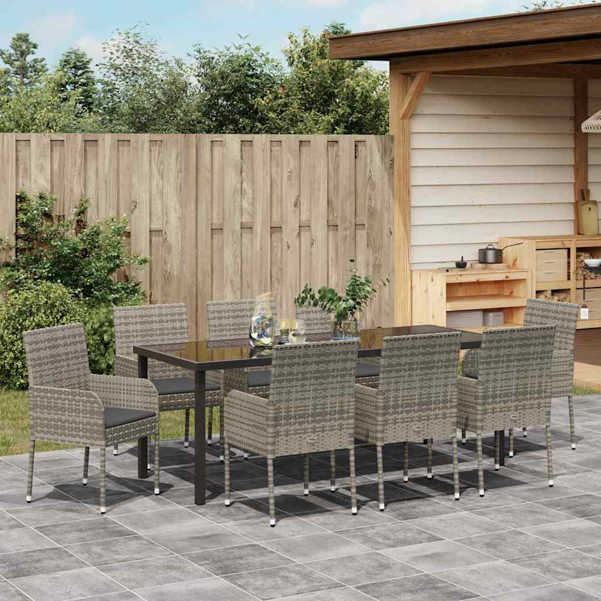 Essgruppe im Freien auf der Terrasse: Tisch, Stühle, Pflanzen, Zaun und Holzkonstruktion im Hintergrund. Wolkenhimmel.