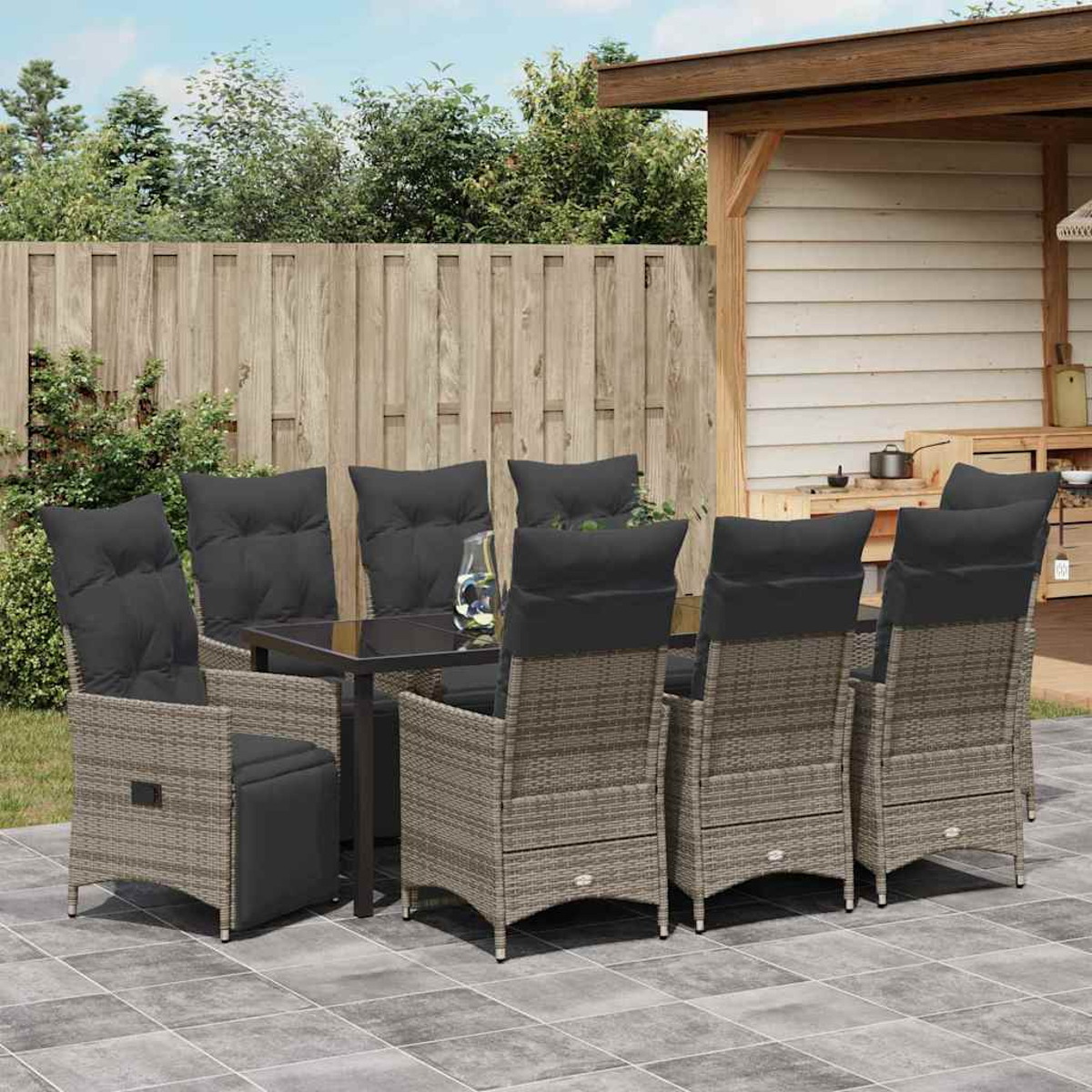 Essgruppe im Freien. Rattan-Stühle mit schwarzen Kissen um einen Tisch mit Glasplatte. Holzzaun und Bäume im Hintergrund.