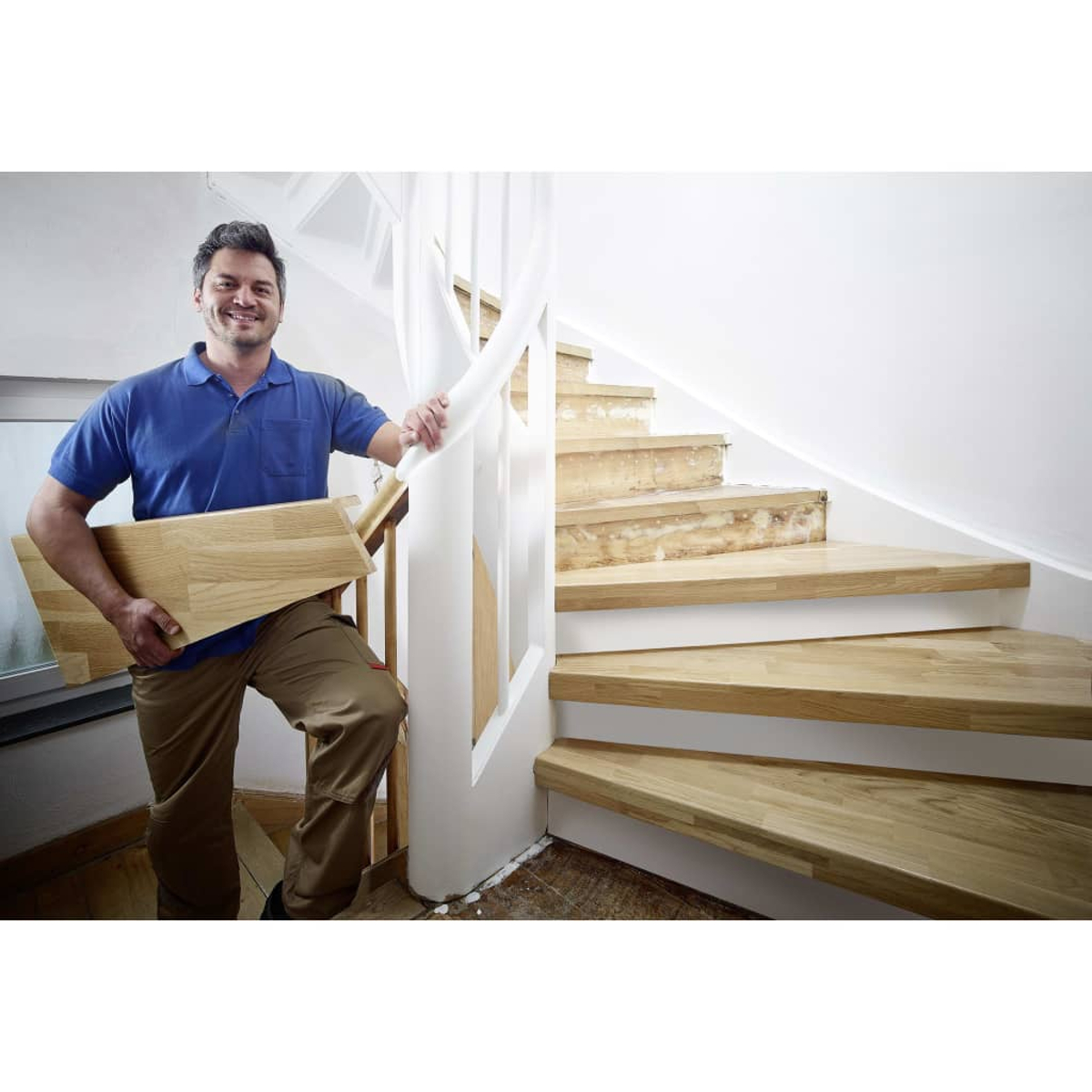 Uomo con assi di legno vicino alle scale. Tiene in mano delle assi, indossa una camicia blu e pantaloni kaki. Scale in legno e finiture bianche.