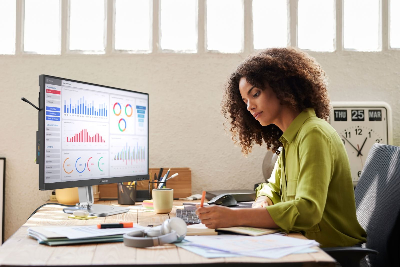 Kobieta siedzi przy biurku, pisząc. Monitor wyświetla wykresy. Zegar i okno w tle.