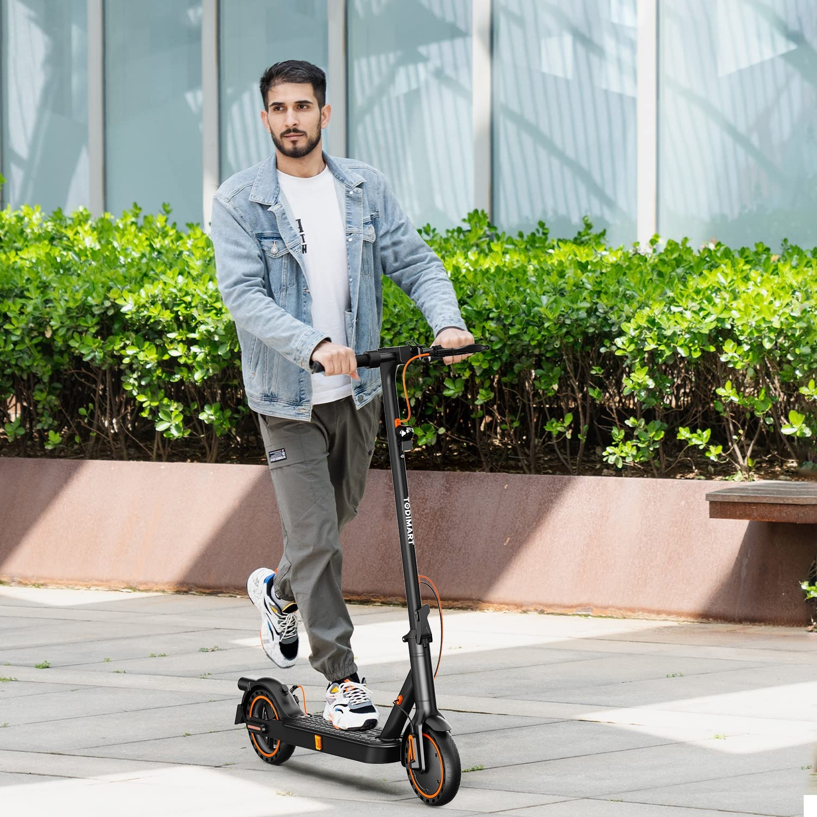 Mężczyzna jedzie na hulajnodze elektrycznej. Ma na sobie dżinsową kurtkę i szare spodnie. Zielone krzewy w tle.