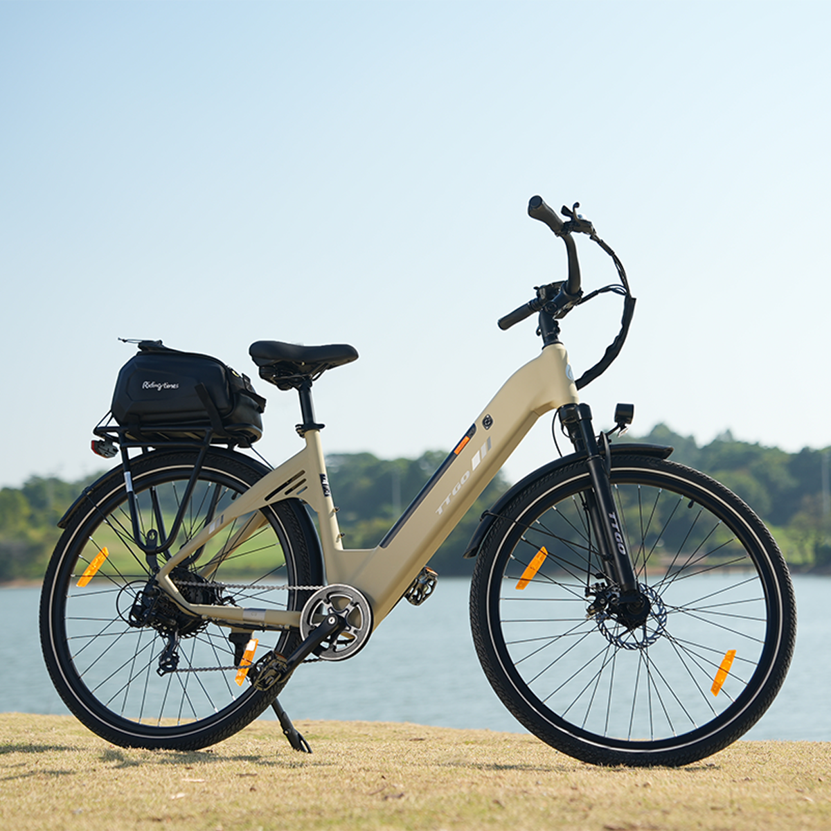 Beżowy rower elektryczny zaparkowany na trawie z jeziorem i drzewami w tle. Z tyłu ma czarną torbę.