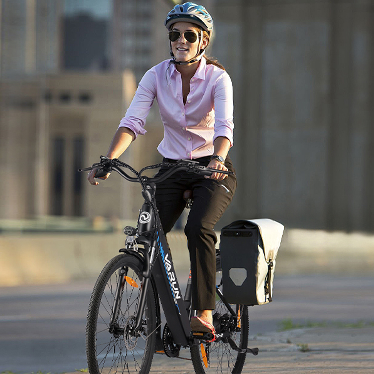 Kobieta jedzie na czarnym rowerze elektrycznym, ubrana w różową koszulę, kask i okulary przeciwsłoneczne. Dołączona jest biała torba.