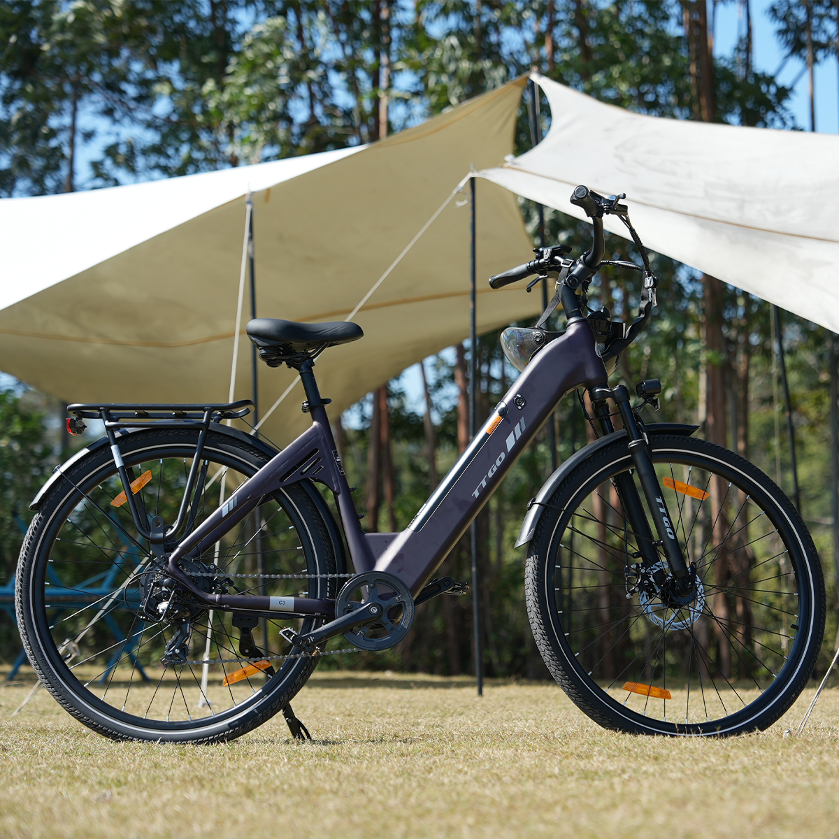 Fioletowy e-bike zaparkowany na trawie. Biały baldachim w tle, z drzewami. Jest słoneczny dzień.