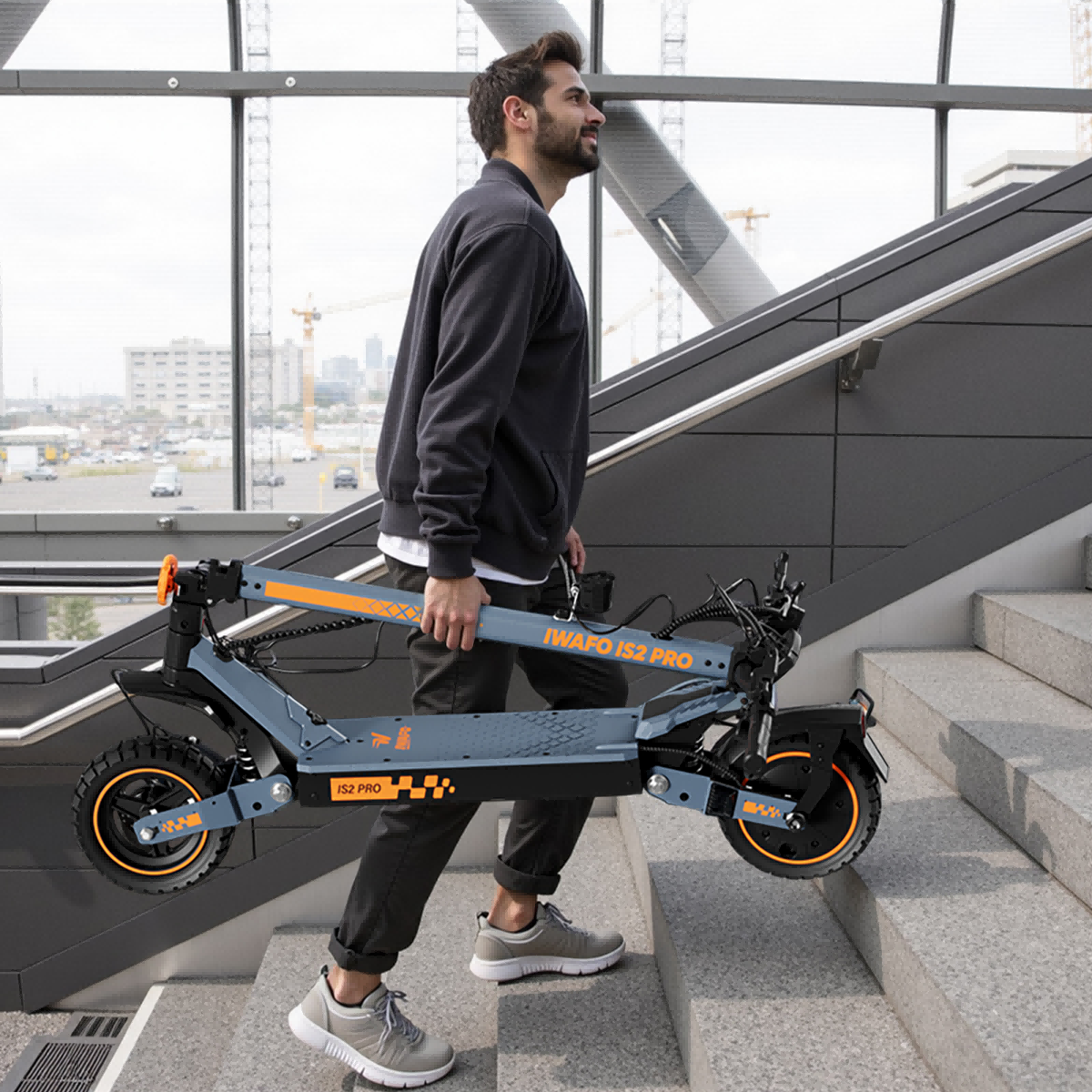 Mężczyzna niesie złożoną szarą hulajnogę po schodach. Hulajnoga ma pomarańczowe akcenty. Tło stanowią budynki, dźwigi i szklane okna.