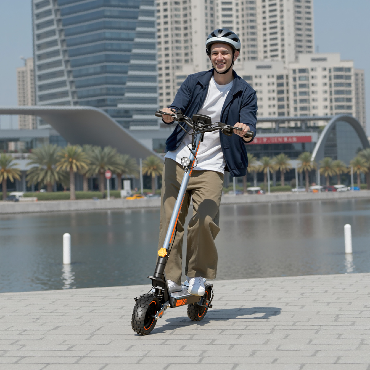 Mężczyzna w kasku jedzie na hulajnodze elektrycznej. W tle budynki miejskie i woda, po bokach palmy.