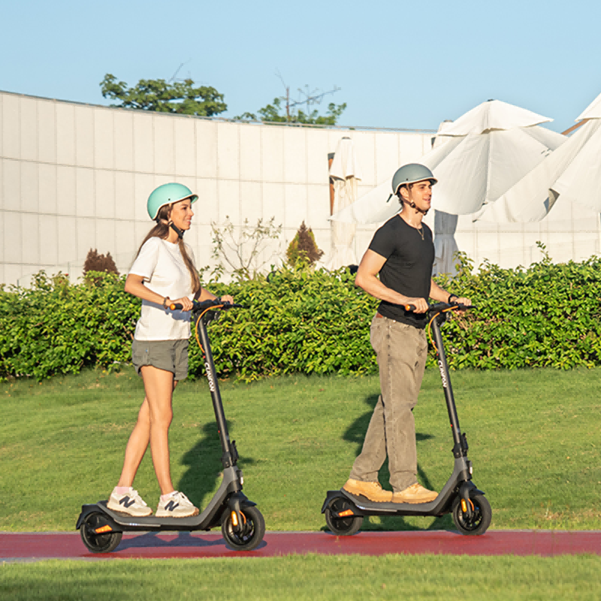 Dwie osoby jadą na hulajnogach po czerwonej ścieżce. Mają kaski. Zielone krzewy i biały budynek w tle.