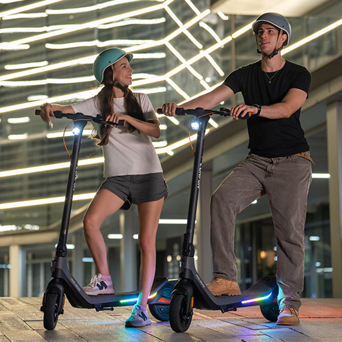 Mężczyzna i kobieta stoją na skuterach. Oboje mają kaski. Tło: oświetlony budynek nocą.