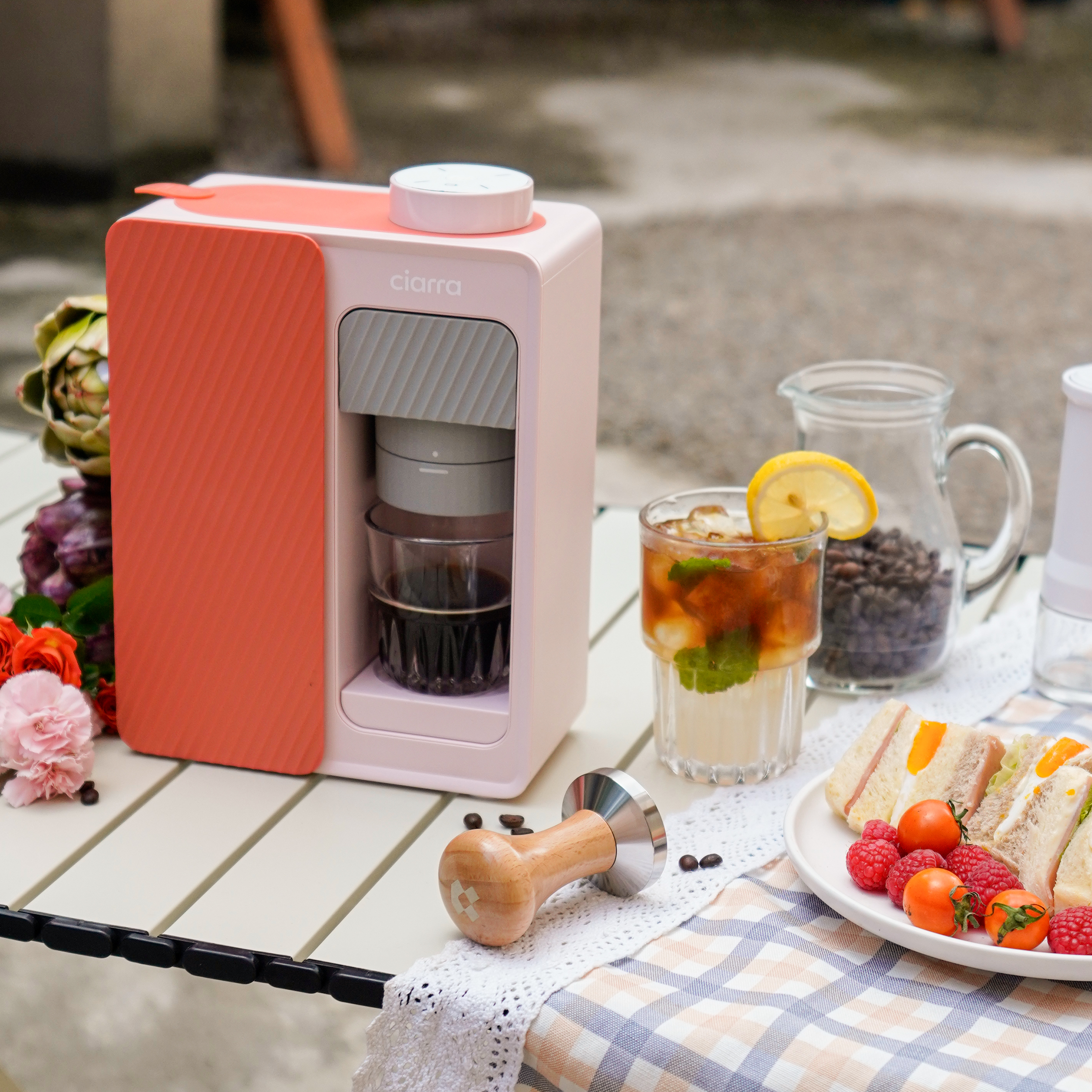 Kaffeemaschine, Eistee, Sandwich und Früchte auf einem Tisch. Blumen und Kaffeebohnen sind in der Nähe.