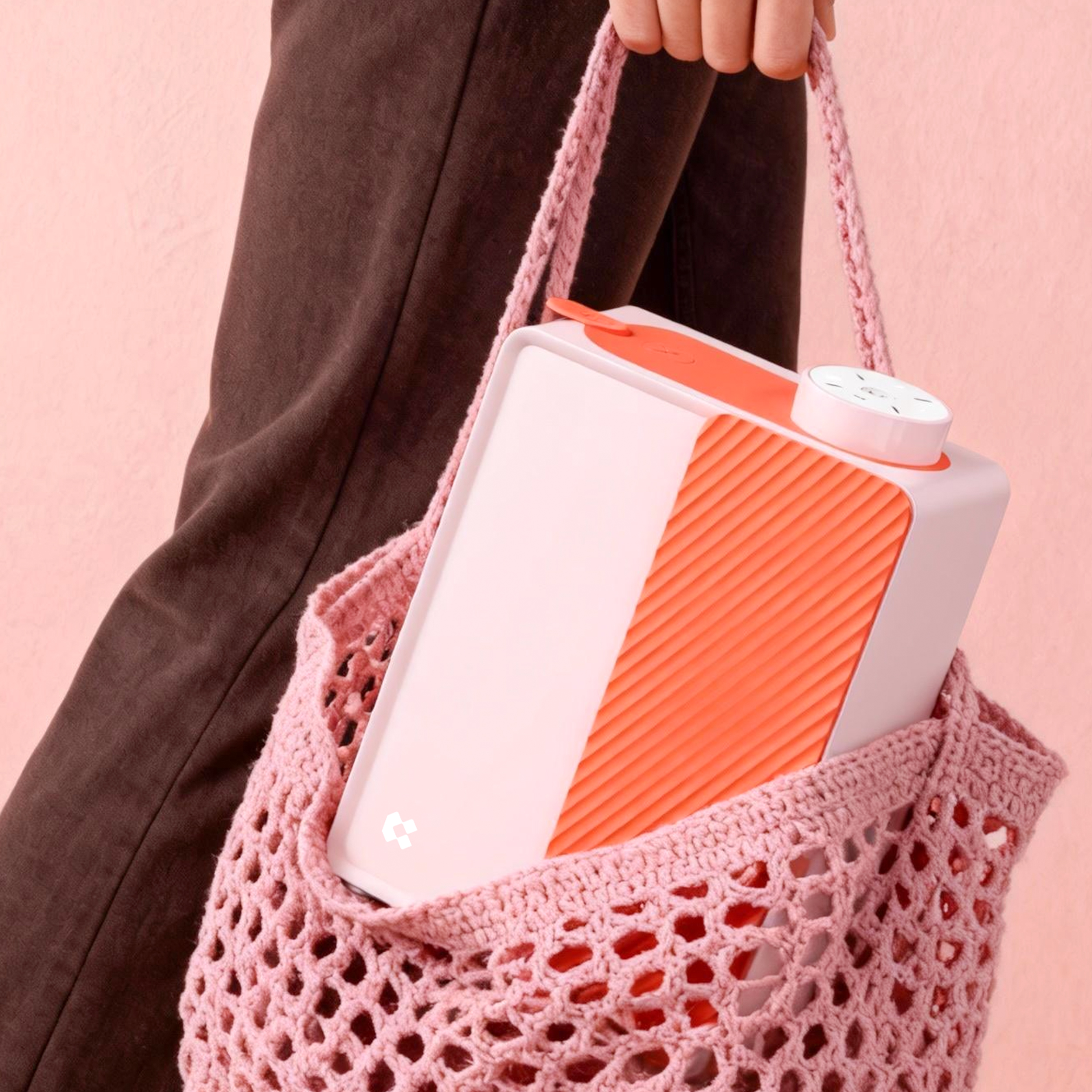 Eine Person hält eine rosa gehäkelte Tasche mit einem weißen und orangefarbenen tragbaren Gerät darin, vor einem rosa Hintergrund.