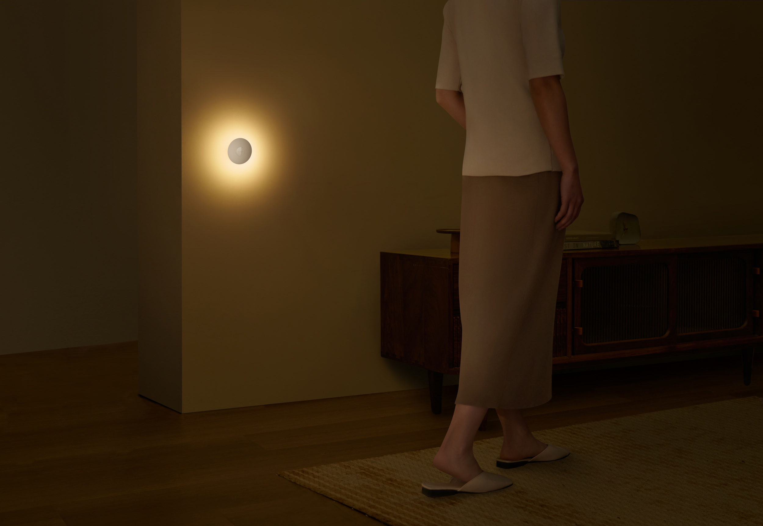 Eine Person in einem Raum, in der Nähe einer Wand mit einem beleuchteten Licht. Ein Holzschrank und ein Teppich sind ebenfalls sichtbar.