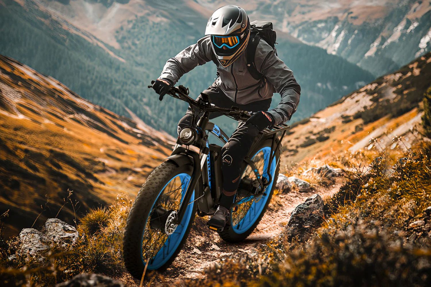 Una persona monta en una bicicleta de montaña negra y azul por un sendero rocoso. Las montañas están al fondo. El ciclista lleva un casco.