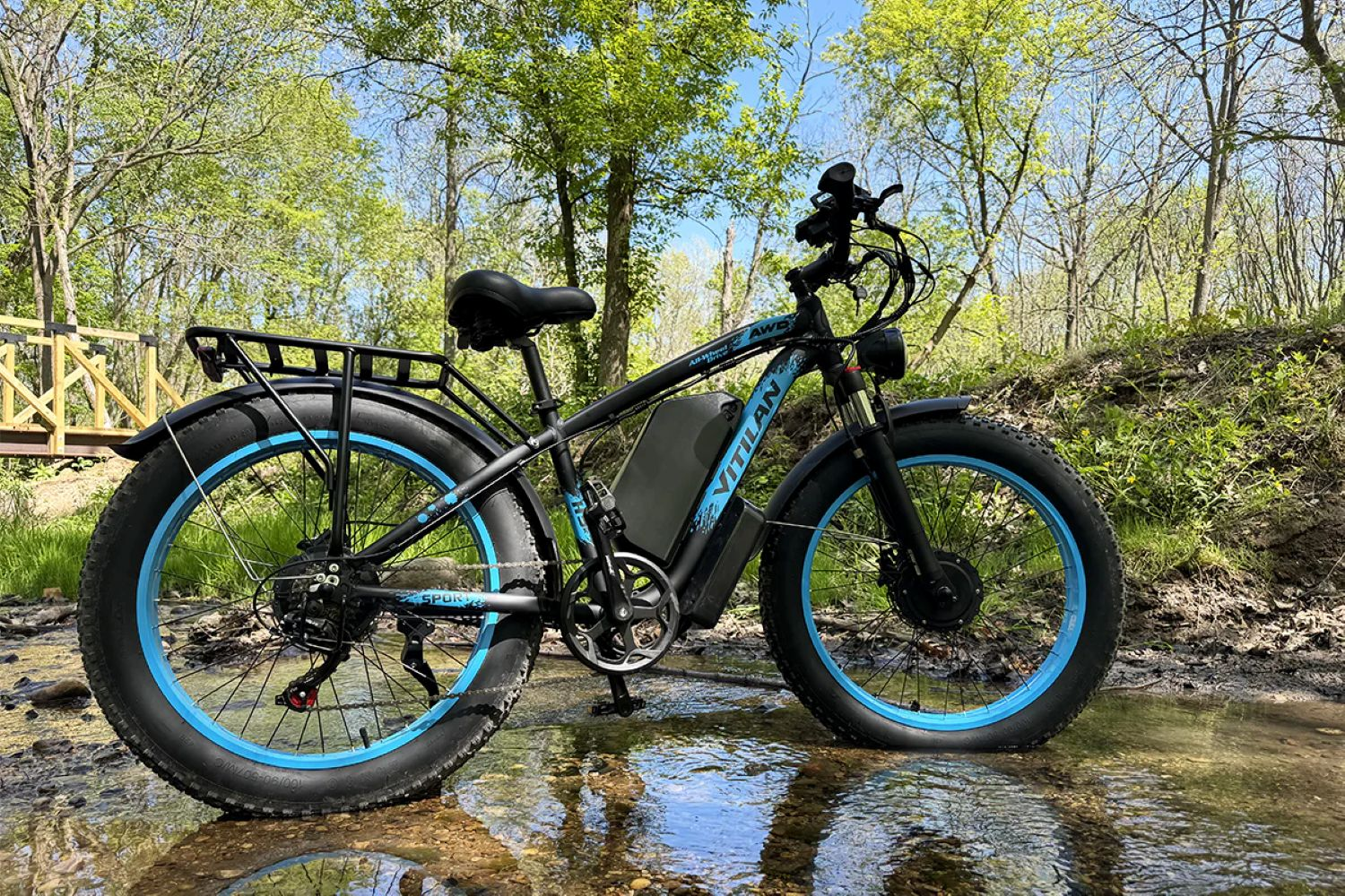 Una e-bike de llanta ancha negra y azul está estacionada en un arroyo poco profundo. Hay árboles al fondo y un puente de madera a la izquierda.