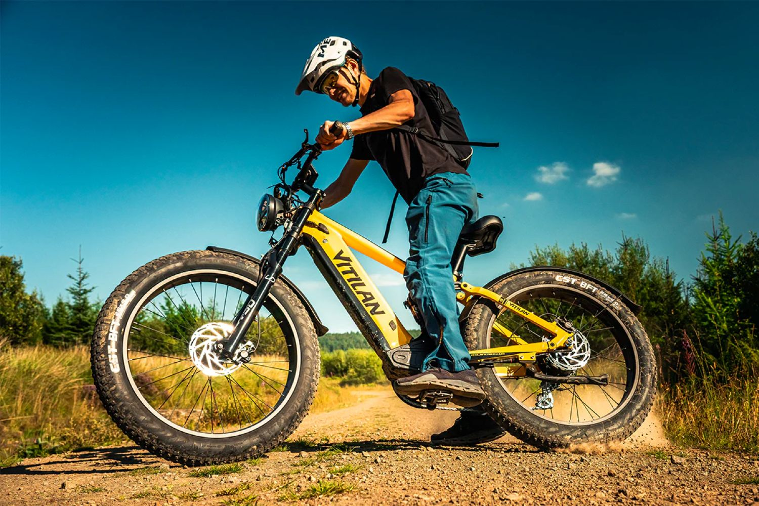 Un hombre monta una bicicleta eléctrica Vitilan amarilla por un camino de tierra. Lleva casco y pantalones azules. Cielo azul claro.