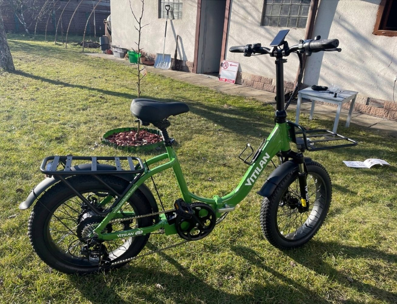 Bicicleta eléctrica Vitilan verde sobre césped. Asiento, neumáticos y portaequipajes negros. Fondo: casa, árbol y herramientas.