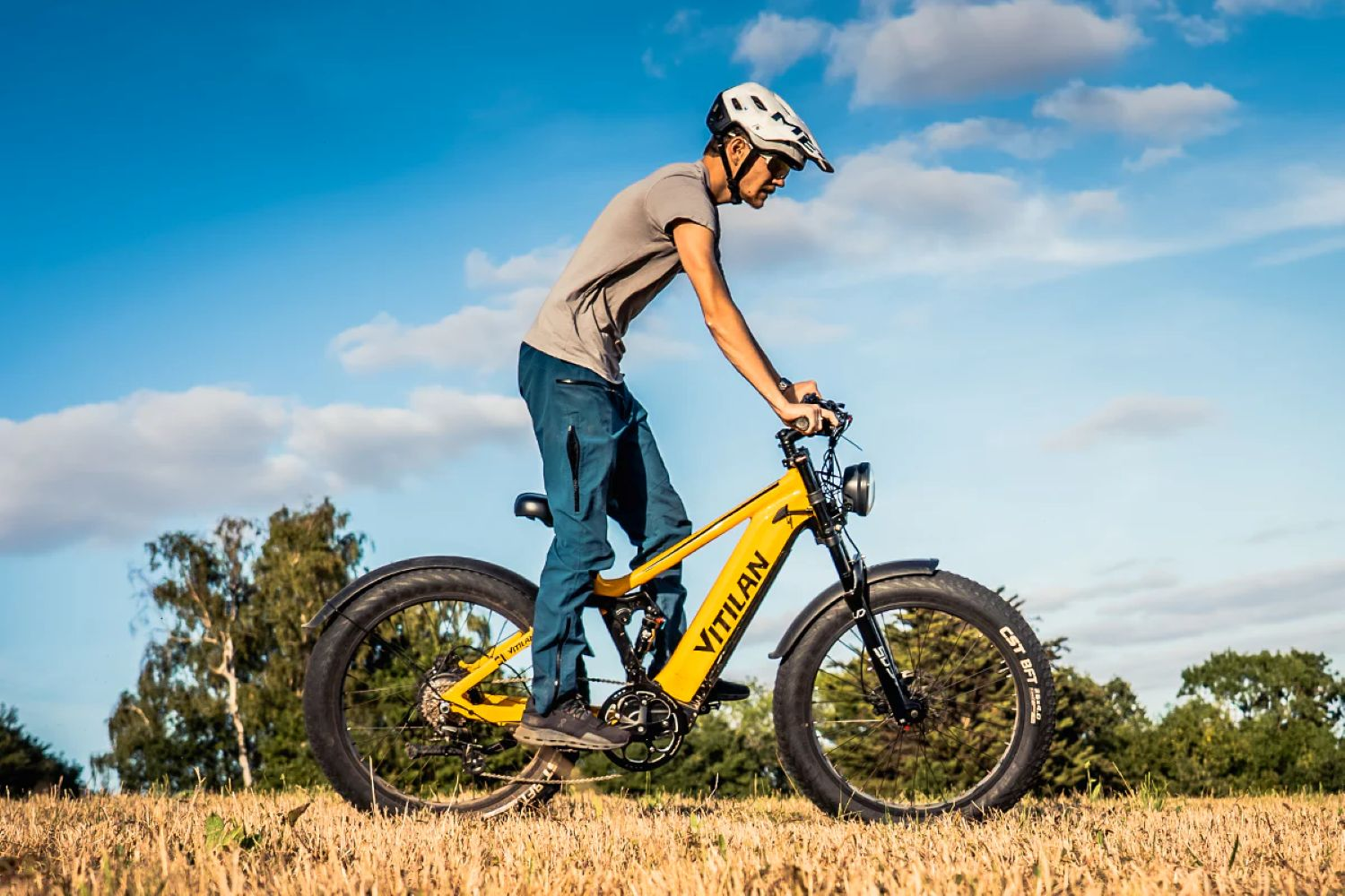 Un hombre monta en una bicicleta eléctrica amarilla en un campo. Lleva casco, camiseta gris y pantalones azules. Cielo azul con nubes.