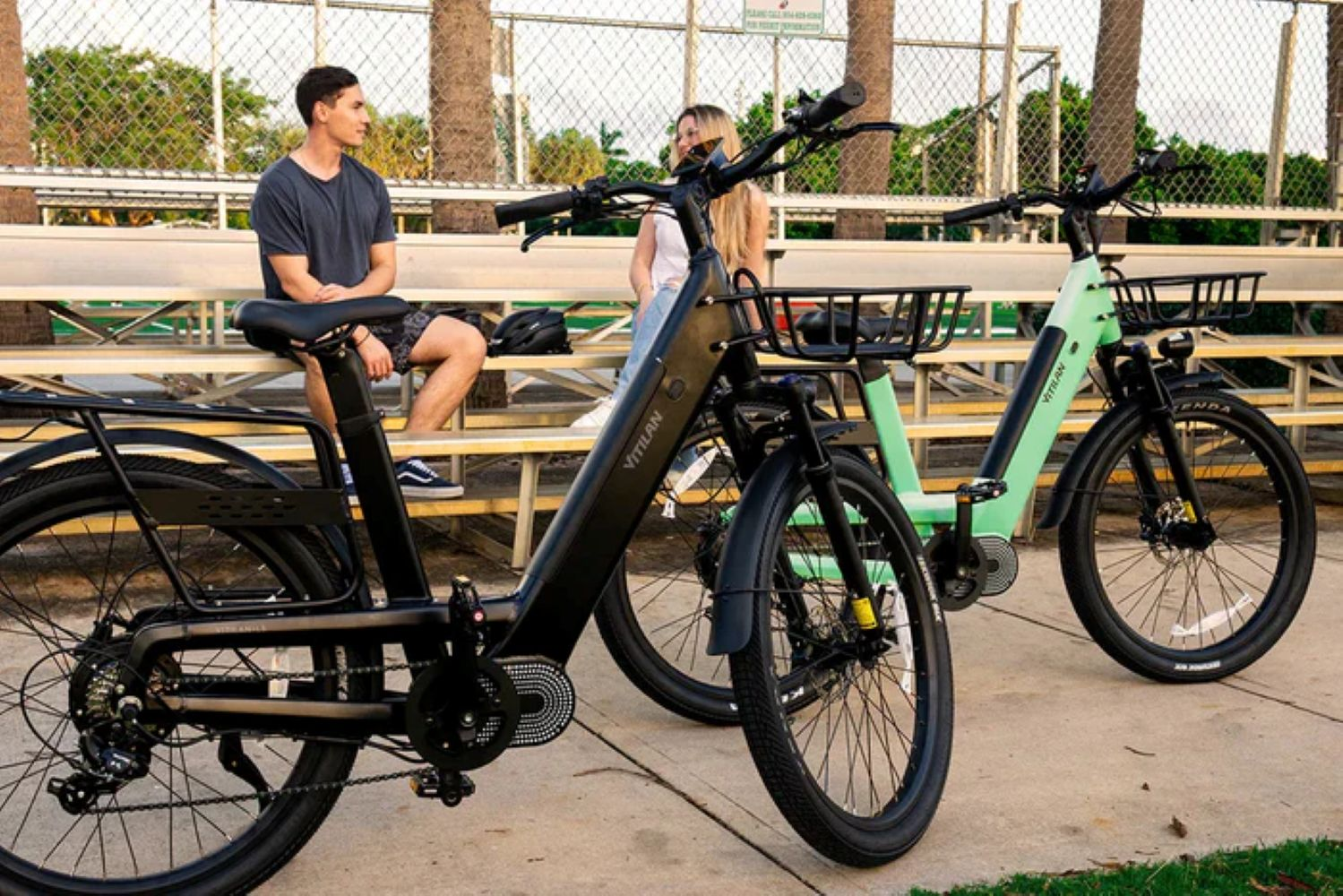 Dos bicicletas con cestas estacionadas frente a gradas. Un hombre y una mujer sentados en las gradas se miran.