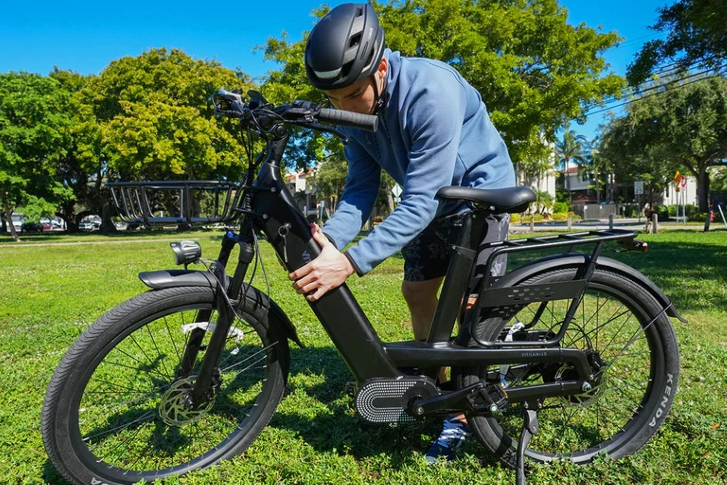 Un hombre con casco ajusta la batería de una bicicleta eléctrica negra. Está sobre hierba, con árboles y cielo azul al fondo.
